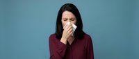 Woman with long dark hair, appearing to be of mixed ethnicity, wearing a burgundy long-sleeved shirt against a blue background, holding tissue to her nose while sneezing or coughing. Useful for healthcare, wellness, and illness-related content.
