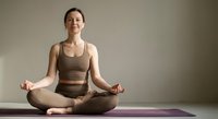 Young Caucasian woman practicing yoga meditation in lotus pose on purple mat wearing brown athletic wear against neutral background for wellness content.