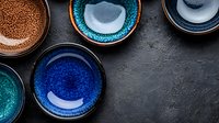 Handcrafted ceramic bowls with vibrant glazed finishes in blue, turquoise and copper tones arranged on dark stone surface, showcasing artisanal pottery craftsmanship.