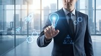 Professional businessman in dark suit touching glowing lightbulb icon with digital innovation symbols in modern office environment for business technology concepts.