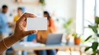 Close-up of hand holding blank white card in modern office with diverse team working in background, suitable for business networking and corporate communication concepts.