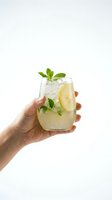 Woman's hand holding a stemless glass with iced lemonade, fresh mint leaves, and lemon slice on a white background.