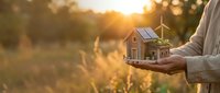 Hands holding miniature eco-friendly house model with solar panels and wind turbine at golden sunset for sustainable energy and green living concepts.