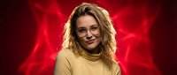 Young blonde woman with curly hair wearing glasses and yellow turtleneck sweater smiling confidently against dramatic red background lighting.
