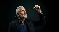 Senior man with gray hair and beard, wearing black blazer and dark blue shirt, holding paintbrush upward against black background. Artist, creative professional, painter concept for design, education, business.