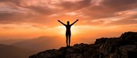 Person with raised arms celebrating success on mountain peak at golden sunset with dramatic orange sky and layered mountain ranges in background.