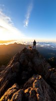 Silhouette of hiker and dog standing on mountain peak at sunrise, dramatic rocky landscape with clouds below and golden morning light illuminating rugged terrain.