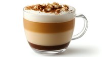 Layered coffee drink in clear glass mug with creamy foam top, caramel middle layer, and chocolate base, sprinkled with cinnamon and nuts.