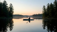 Silhouette of person kayaking on calm lake at sunset with pine forest reflection creating peaceful outdoor recreation scene for adventure and nature enthusiasts.