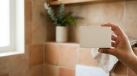 Person holding blank white card in modern bathroom with terracotta tiles, potted plant, and natural light for business, greeting, or promotional messaging.