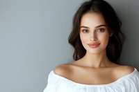 Young attractive brunette woman with natural makeup wearing white off-shoulder top smiling at camera against gray background for beauty and lifestyle content.