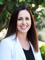 Young Caucasian businesswoman with long brown hair wearing white blazer and black top smiling confidently against blurred natural background. Professional headshot portrait.