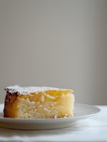 Slice of homemade lemon cake dusted with powdered sugar on white plate against neutral background, showcasing moist texture and bright citrus filling.
