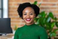 Young African American businesswoman with natural curly hair and bright red lipstick wearing green sweater smiling confidently in modern office environment.