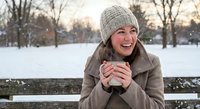 Young Caucasian woman in beige coat and knit hat enjoying hot drink outdoors in snowy winter park, smiling warmly while holding steaming mug on cold day.