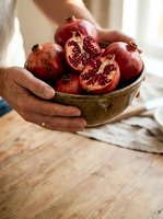 Hands holding ceramic bowl filled with fresh ripe pomegranates, some cut open showing seeds, on wooden table with natural light for food and lifestyle content.