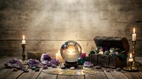 Fortune teller mystic table with crystal ball, tarot cards, amethyst crystals, treasure chest and burning candles on rustic wooden background.