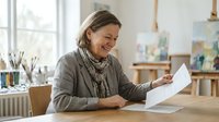 Middle-aged woman with light brown hair and fair complexion, wearing casual gray cardigan and patterned scarf, smiling while reviewing documents at wooden desk in bright studio workspace with art supplies and easels.