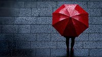Silhouette of person holding bright red umbrella against dark rainy background with water drops on pavement, creating dramatic contrast and moody atmosphere.