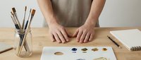 Artist hands working with watercolor paints on wooden table in creative studio workspace with brushes and palette for artistic projects.