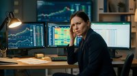 Focused female financial analyst in dark blazer analyzing stock market data on multiple computer monitors displaying trading charts and graphs in modern office workspace.