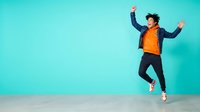Young male, South Asian ethnicity, wearing yellow shirt and dark jacket, jumping with arms raised in celebration against turquoise studio background. Perfect for lifestyle, success, happiness, and youth-oriented marketing campaigns.