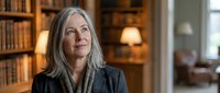 Mature woman with long gray hair wearing dark blazer in library setting with bookshelves and warm lighting, suitable for business, education, and professional portraits.