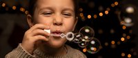 Young Caucasian boy in cozy sweater blowing soap bubbles with warm bokeh lights creating magical atmosphere for children activities and play concepts.