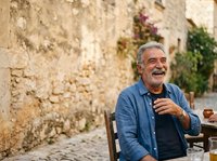 Senior man with gray hair and Mediterranean ethnicity, wearing casual blue shirt, seated outdoors against weathered stone wall with ivy. Suitable for lifestyle, travel, and aging themes.