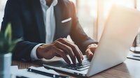 Businessman in formal suit working on laptop at wooden desk with natural light, showcasing professional environment for corporate projects.