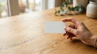 Hand holding blank white business card on wooden table with natural window light and potted plant in background for branding mockup.