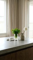 Minimalist home interior with fresh green plant in glass vase, ceramic pottery and white stones on marble countertop near bright windows with beige curtains.