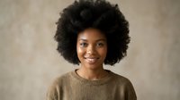 Young African American woman with natural afro hairstyle smiling warmly in cozy brown sweater against neutral textured background for lifestyle content.