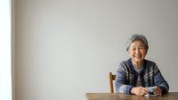 Senior Asian woman with gray hair smiling at wooden table against white wall background, wearing dark patterned cardigan, suitable for lifestyle and portrait use.