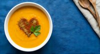 Creamy orange soup with heart-shaped spice garnish and fresh herbs in white bowl on blue textured background with wooden spoons and linen napkin.