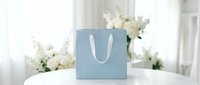 Blue shopping bag with white handles on white table with white flowers in background, suitable for retail, e-commerce, and gift packaging designs.