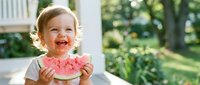 Toddler girl with blonde hair eating fresh watermelon slice outdoors in summer garden setting with natural sunlight and green foliage background.