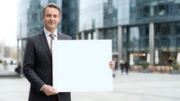 Professional Caucasian businessman in dark suit holding blank white sign outdoors in modern urban business district for corporate presentations.