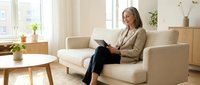 Professional mature businesswoman using tablet computer while relaxing on modern sofa in bright contemporary living room interior for remote work.