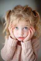 Young girl with blonde hair and striking blue eyes wearing cozy pink knit sweater rests face in hands with dreamy expression, soft natural lighting creates gentle mood.