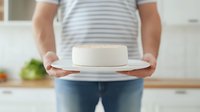 Caucasian person in striped shirt holding white ceramic plate with fresh unfrosted round cake in bright modern kitchen, focus on dessert, home baking concept.