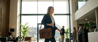 Professional businesswoman walking through modern office lobby with leather briefcase during daytime meeting preparation.