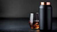Black matte insulated water bottle with copper lid next to crystal glass filled with whiskey on dark background, minimalist drink composition.