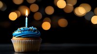 Single blue frosted cupcake with lit birthday candle against dark background with golden bokeh lights creating festive celebration atmosphere.