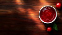 Red cherry sauce in white ceramic bowl on dark wooden background with fresh berries and green leaves, top view with copy space for menu design.