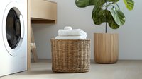 Clean folded white towels in round woven rattan basket near washing machine and houseplant in modern minimalist laundry room interior with wooden elements.