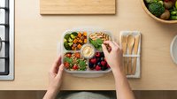 Overhead view of hands holding a compartmentalized meal prep container filled with fresh vegetables, grains, and berries on wooden table. Healthy eating, nutrition planning.