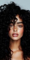 Young woman with natural freckles and voluminous curly black hair gazing at camera. Soft makeup highlighting full lips and glowing skin creates modern beauty portrait.