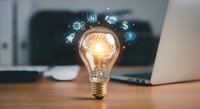 Glowing light bulb with digital business icons floating above on wooden desk with laptop in modern office workspace for innovation and creativity concepts.