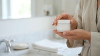 Middle-aged woman with light skin tone holding white cosmetic jar in modern bathroom setting, showcasing skincare product packaging for beauty and wellness marketing.